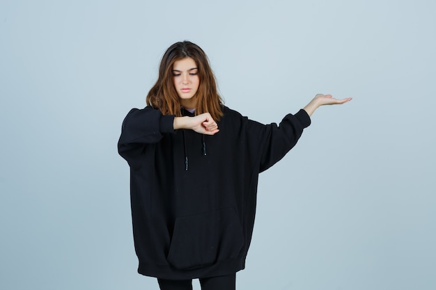 Jonge dame die pols bekijkt terwijl iets in oversized hoodie, broek wordt getoond en vergeetachtig kijkt. vooraanzicht.