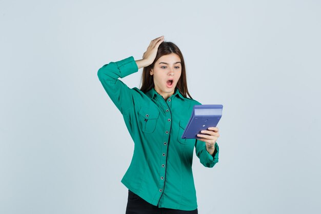 Jonge dame die rekenmachine bekijkt terwijl hand op hoofd in groen overhemd wordt gehouden en geschokt, vooraanzicht kijkt.