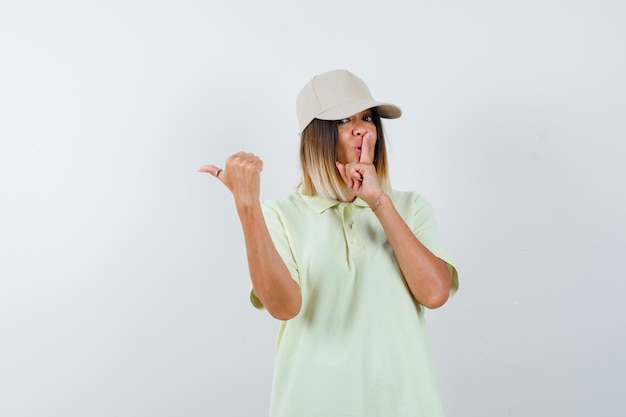 Jonge dame die stilte gebaar toont terwijl ze naar de linkerkant wijst met duim in t-shirt, pet en er zelfverzekerd uitziet, vooraanzicht.