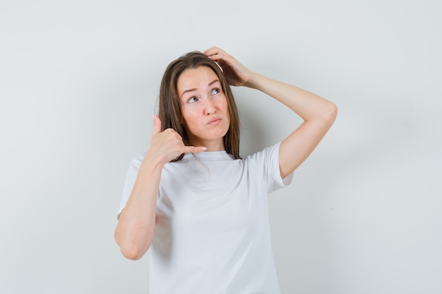 Jonge dame die telefoongebaar toont terwijl het hoofd in wit t-shirt krabt en vergeetachtig kijkt