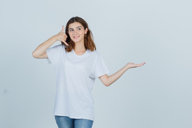 Jonge dame die telefoongebaar toont terwijl ze doet alsof ze iets in t-shirt, spijkerbroek vasthoudt en er zelfverzekerd uitziet, vooraanzicht.