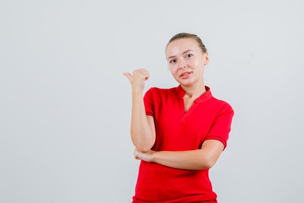 Jonge dame die terug met duim in rood t-shirt wijst en vrolijk kijkt