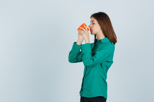 Jonge dame die thee drinkt uit oranje kop in overhemd en gericht, vooraanzicht kijkt.