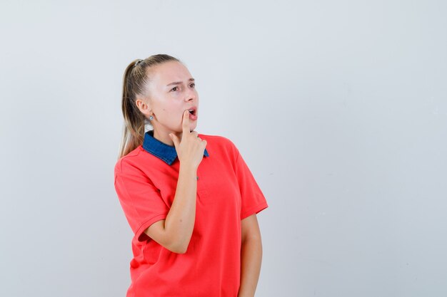 Jonge dame die vinger dichtbij mond in t-shirt houdt en peinzend kijkt. vooraanzicht.