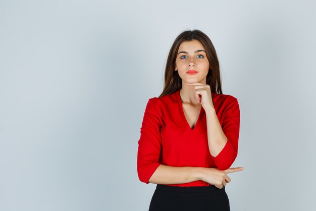 Jonge dame die vinger onder kin in rode blouse, rok houdt en mooi kijkt