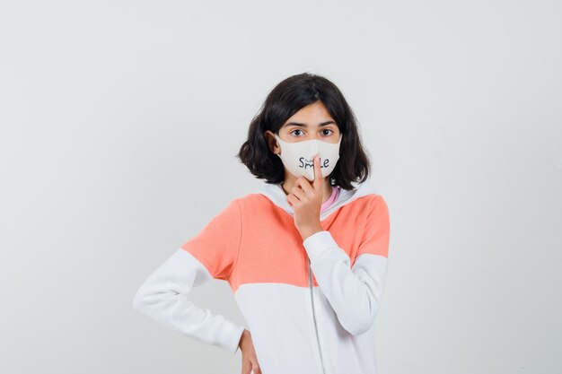 Jonge dame die vinger op haar masker in hoodie, gezichtsmasker houdt en kalm kijkt.