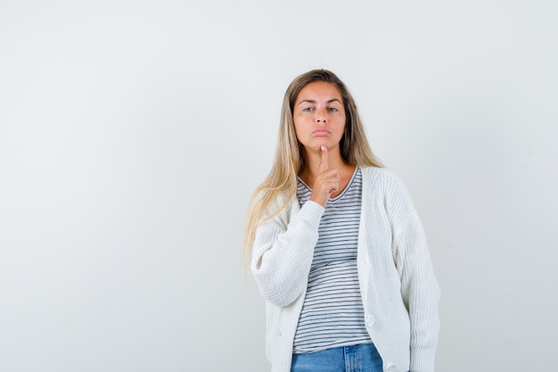 Jonge dame die vinger op kin in t-shirt, jasje, spijkerbroek houdt en gekwetst, vooraanzicht kijkt.