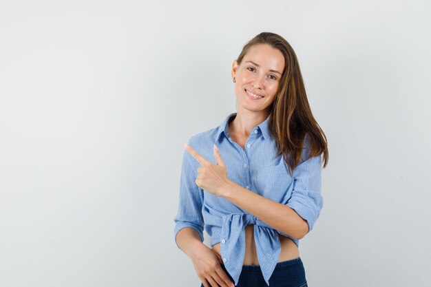 Jonge dame die weg wijst in blauw overhemd, broek en vrolijk kijkt.