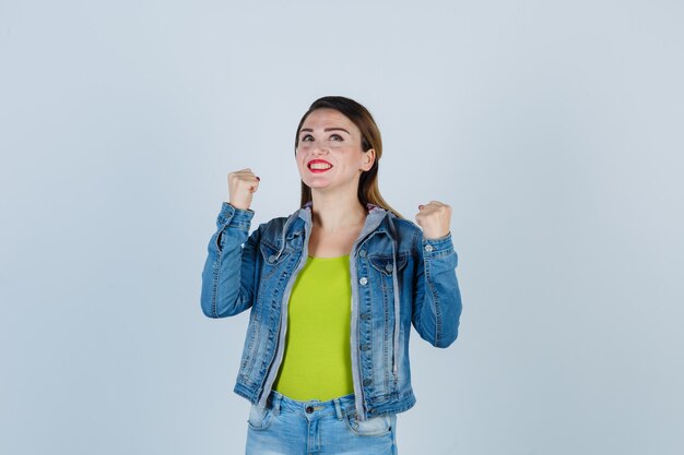 Jonge dame die winnaargebaar in denimoutfit toont en er vrolijk uitziet, vooraanzicht.