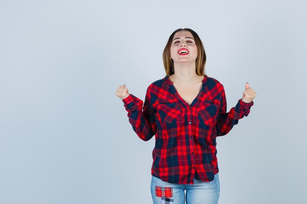 Jonge dame die winnaargebaar toont in geruit overhemd, jeans en opgetogen kijkt, vooraanzicht.