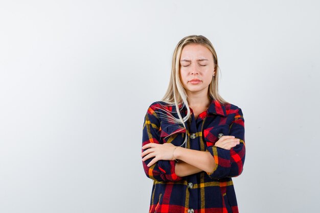 Jonge dame die zich met gekruiste armen in geruit overhemd bevindt en weemoedig kijkt. vooraanzicht.