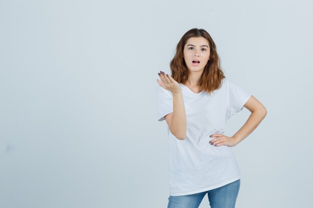 Jonge dame die zich uitstrekt hand in vragend gebaar in t-shirt, spijkerbroek en op zoek verbaasd, vooraanzicht.