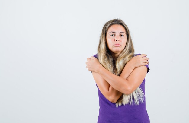 Jonge dame die zichzelf in violet t-shirt knuffelt en triest, vooraanzicht kijkt.