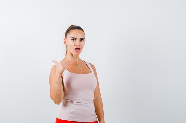 Jonge dame duimen omhoog in beige tanktop en kijkt verbaasd, vooraanzicht.