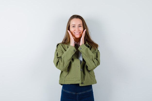 Jonge dame hand in hand in de buurt van gezicht in shirt, jas en ziet er heerlijk uit, vooraanzicht.