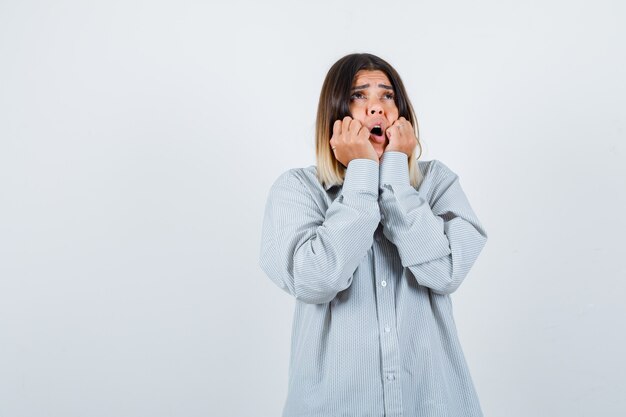 Jonge dame hand in hand op de wangen terwijl ze opkijkt in een oversized shirt en verbijsterd kijkt, vooraanzicht.