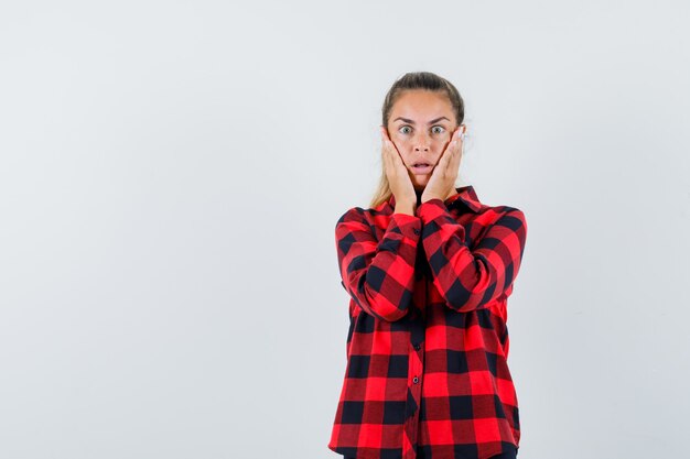 Jonge dame hand in hand op wangen in casual shirt en verbaasd op zoek. vooraanzicht.