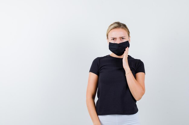 Jonge dame houdt hand op masker in t-shirt, broek, medisch masker en ziet er serieus uit