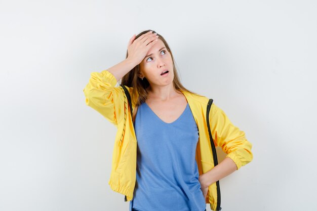 Jonge dame houdt hand op voorhoofd in t-shirt, jas en kijkt peinzend, vooraanzicht.