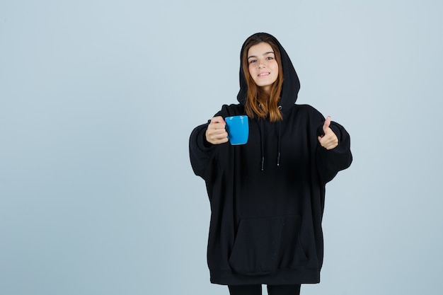 Jonge dame houdt kopje terwijl ze dreun in oversized hoodie, broek en ziet er mooi uit, vooraanzicht.