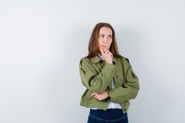 Jonge dame houdt vinger op kin in blouse, jas en kijkt peinzend, vooraanzicht.