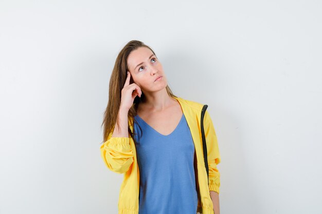 Jonge dame houdt vinger op tempels in t-shirt, jas en kijkt peinzend. vooraanzicht.