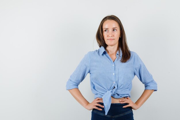 Jonge dame in blauw shirt, broek die weg met handen op taille kijkt en peinzend kijkt