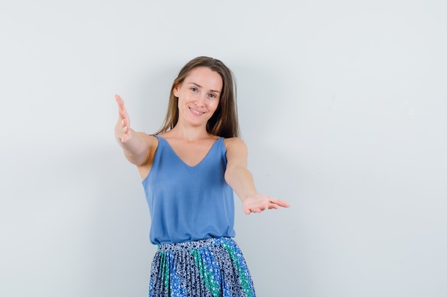 Jonge dame in blauwe blouse, rok die armen opheft om iemand te knuffelen en oprecht, vooraanzicht te kijken.