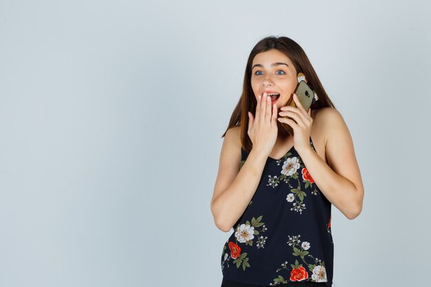 Jonge dame in blouse die op mobiele telefoon praat, hand op mond houdt en zich afvraagt, vooraanzicht.