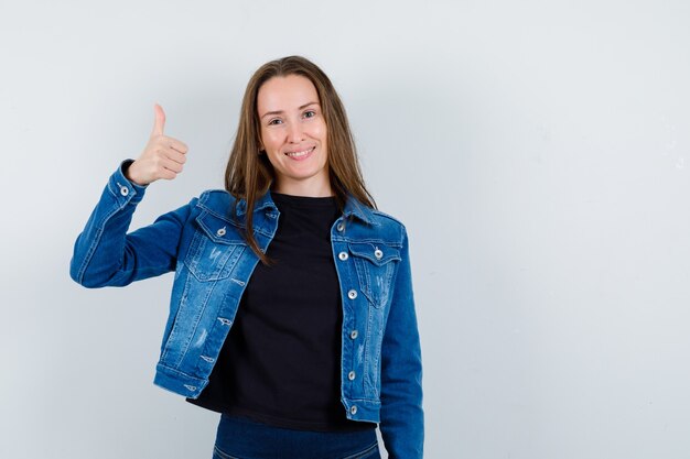 Jonge dame in blouse, jas met duim omhoog en vrolijk, vooraanzicht.