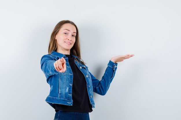 Jonge dame in blouse, jas wijzend op camera, iets laten zien en zelfverzekerd kijken, vooraanzicht.