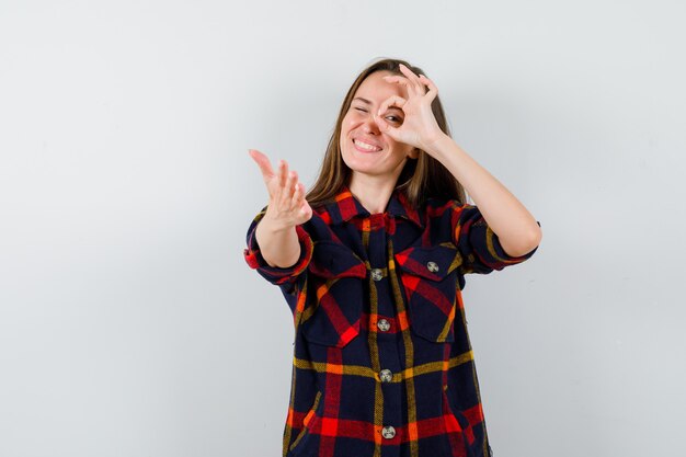 Jonge dame in casual shirt met ok teken op oog, hand uitstrekt en op zoek vrolijk, vooraanzicht.