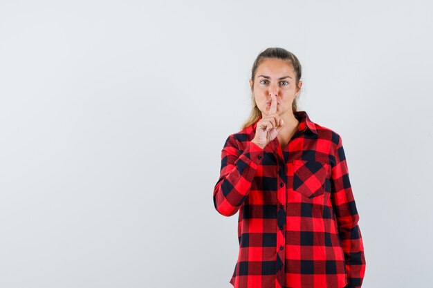 Jonge dame in casual shirt met stilte gebaar en op zoek naar zelfverzekerd, vooraanzicht.