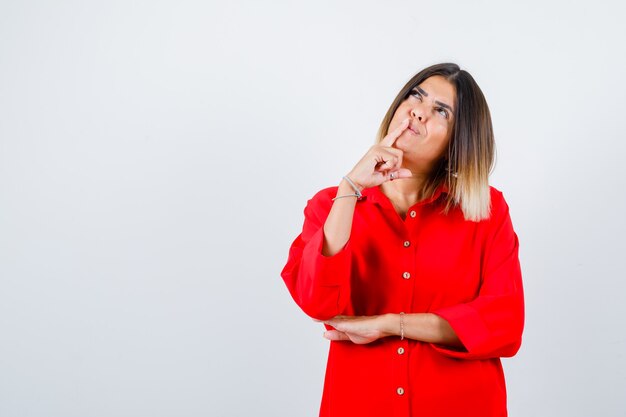 Jonge dame in een rood oversized shirt die de vinger op de mond houdt en er attent uitziet, vooraanzicht.