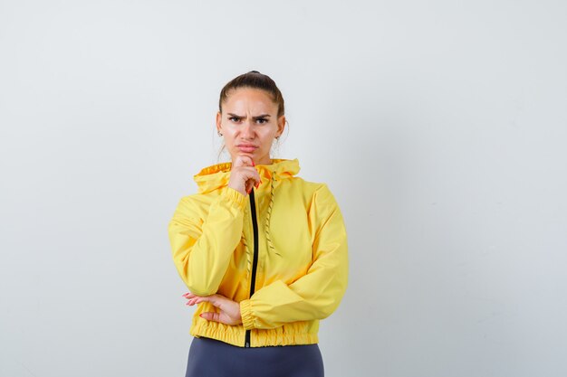 Jonge dame in gele jas die de kin bij de hand steekt en er serieus uitziet, vooraanzicht.