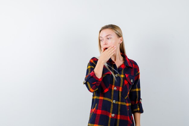 Jonge dame in geruit overhemd die hand op mond houdt terwijl wegkijkend en verbaasd, vooraanzicht kijkt.