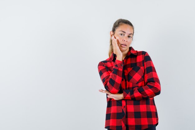 Jonge dame in geruit overhemd die huid op haar wang aanraakt en gracieus kijkt