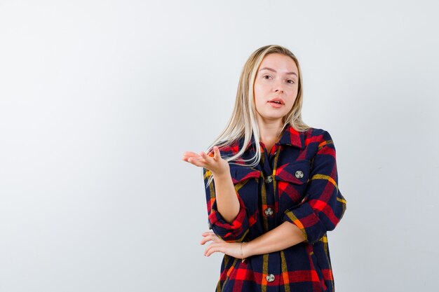 Jonge dame in ingecheckte overhemd strekken hand in vragend gebaar en op zoek hulpeloos, vooraanzicht.