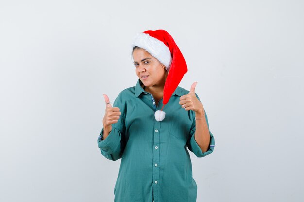 Jonge dame in kerstmuts, shirt duimen opdagen en tevreden kijken, vooraanzicht.