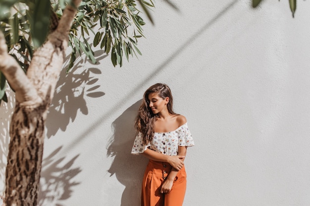 Jonge dame in oranje broek en blouse met blote schouders glimlacht zoet, leunend op witte muur onder boom