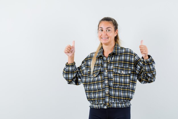 Jonge dame in overhemd, korte broek die dubbele duimen toont en gelukkig, vooraanzicht kijkt.