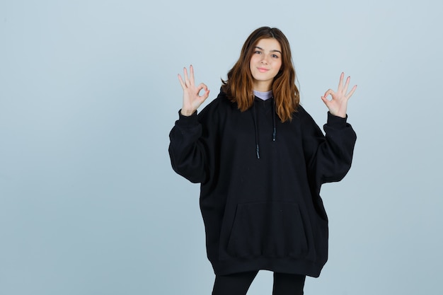 Jonge dame in oversized hoodie, broek die ok gebaar toont en zalig, vooraanzicht kijkt.