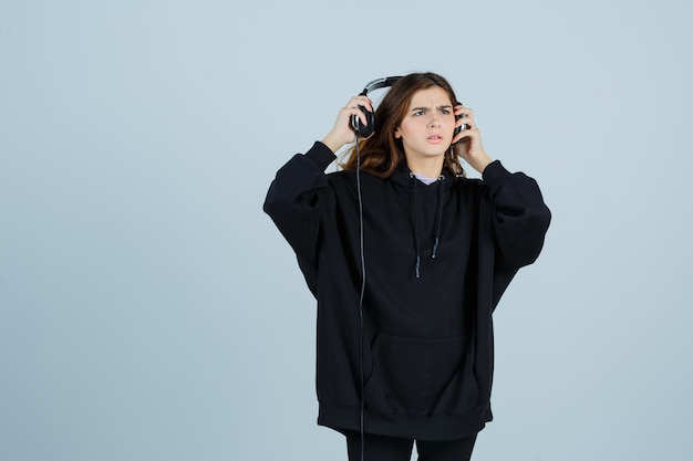 Jonge dame in oversized hoodie, broek met handphones en verbaasd op zoek, vooraanzicht.