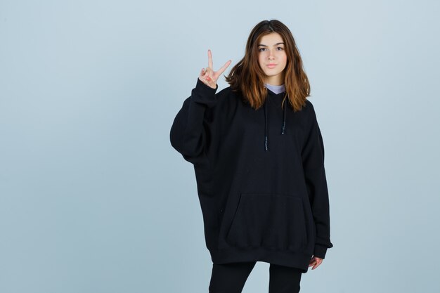 Jonge dame in oversized hoodie, broek met overwinningsteken en op zoek naar geluk, vooraanzicht.