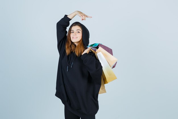 Jonge dame in oversized hoodie, broek met pakjes op de schouder terwijl ze naar de rechterkant wijst en er prachtig uitziet, vooraanzicht.