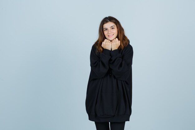 Jonge dame in oversized hoodie, broek met vuisten onder de kin en ziet er schattig uit, vooraanzicht.