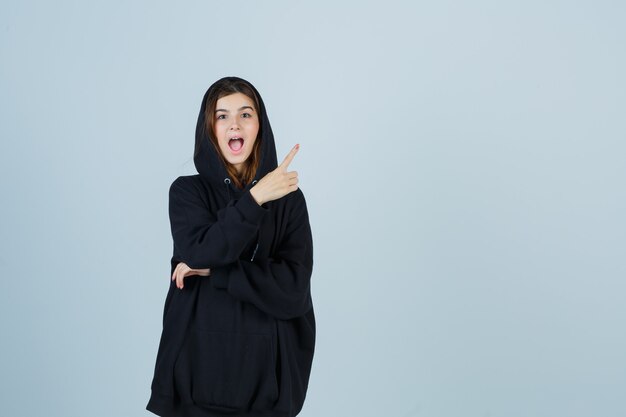 Jonge dame in oversized hoodie, broek wijst naar de rechterbovenhoek en kijkt verbaasd, vooraanzicht.