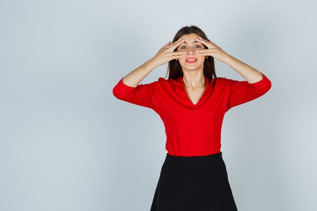 Jonge dame in rode blouse, rok die door vingers kijkt en er schattig uitziet