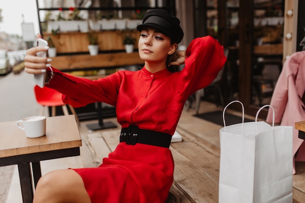 Jonge dame in rode fluweel jurk en met boodschappenzakken, zit in een café en neemt een selfie van haar nieuwe smartphone