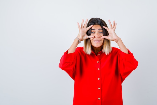 Jonge dame in rood oversized shirt die ogen opent met vingers en er vrolijk uitziet, vooraanzicht.